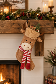 A plush reindeer Christmas stocking with a red and white striped scarf and small gift box, hanging from a rustic wooden mantel decorated with evergreen garland, pinecones, and candles.