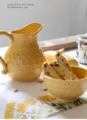 A yellow honeycomb-textured ceramic pitcher and matching bowl with biscotti, styled on a floral table runner with a neutral background.