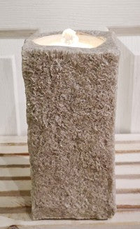 A rectangular stone pillar candle holder with a textured, rough-hewn appearance, filled with a white tea light candle, set against a light-colored paneled background.