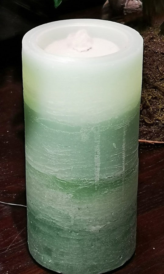 A cylindrical pillar candle with a white wax top and a gradient body that fades from off-white at the top to light green at the bottom, shown on a dark wooden surface.