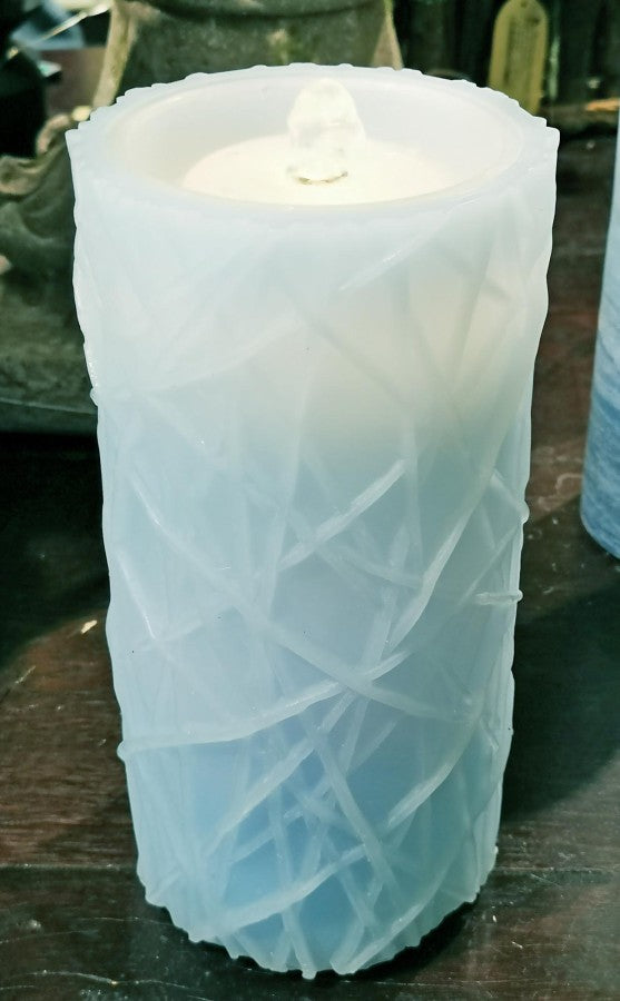 A textured white pillar candle with a crisscross geometric pattern, shown lit on a dark wooden surface.