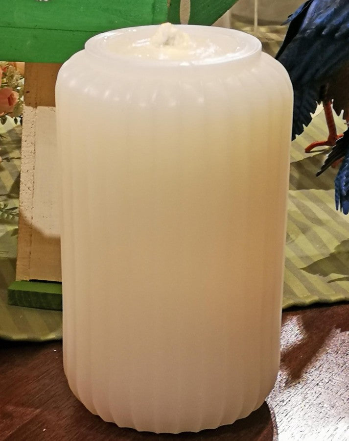 A ribbed, cylindrical white candle with a wick on top, photographed on a wooden surface with a green and striped fabric background.