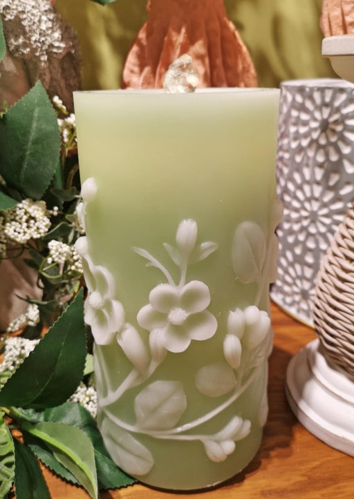 Decorative candle with floral embossing on a wooden surface with greenery and another candle in the background.