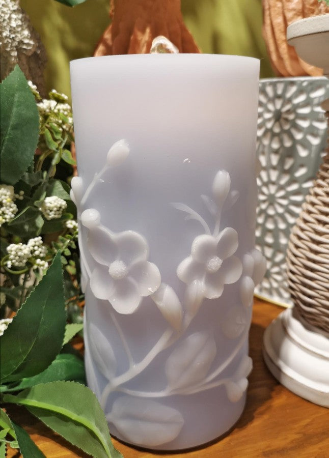 A frosted white pillar candle with embossed white cherry blossom flowers and leaves, displayed on a wooden surface with decorative greenery and other home decor in the background.