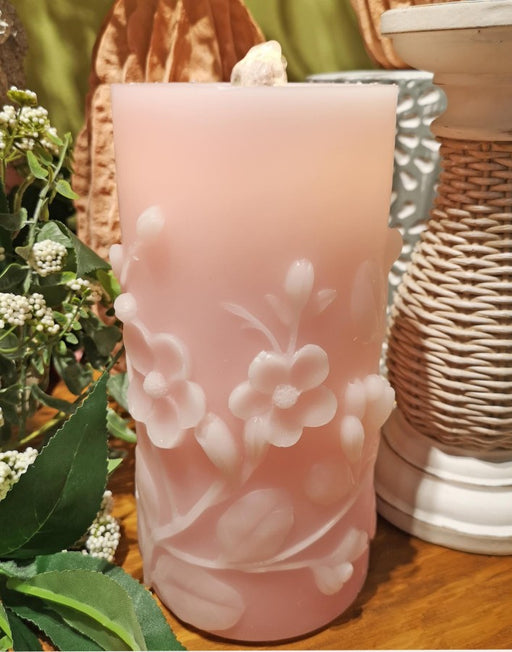Pink candle with floral design on a wooden surface with plants and woven baskets in the background