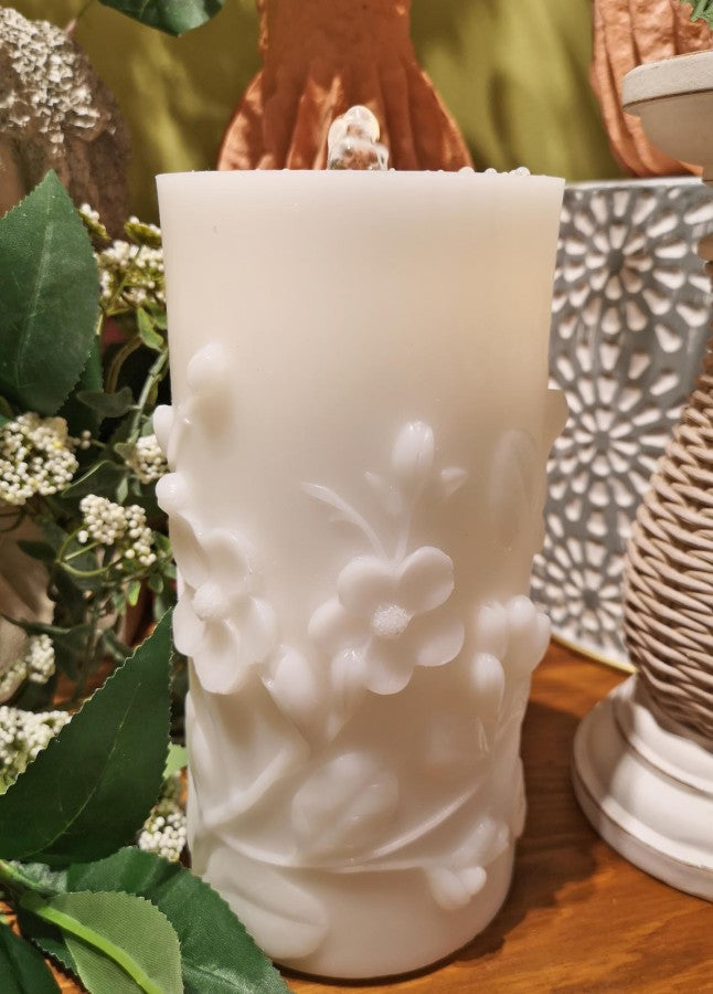Decorative white candle with floral embossing on a wooden surface with greenery.