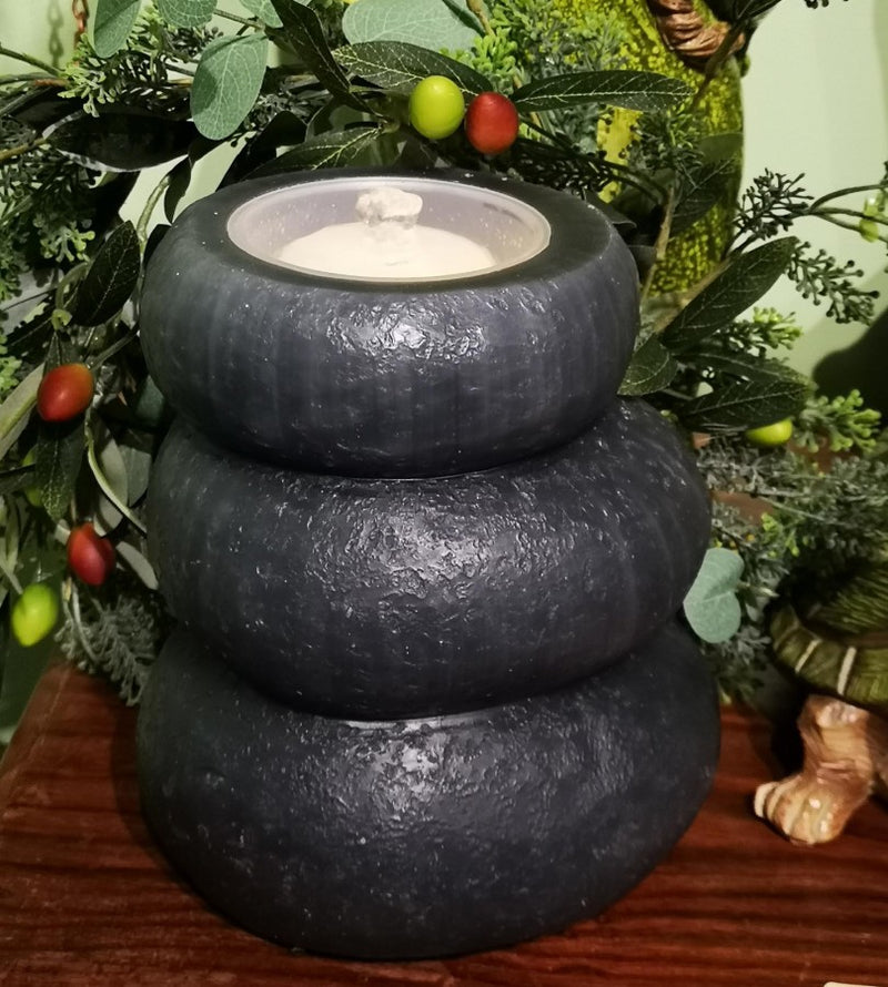 A three-tiered dark gray stone candle with a white wax candle inside, styled on a wooden surface with green foliage and small red berries in the background.