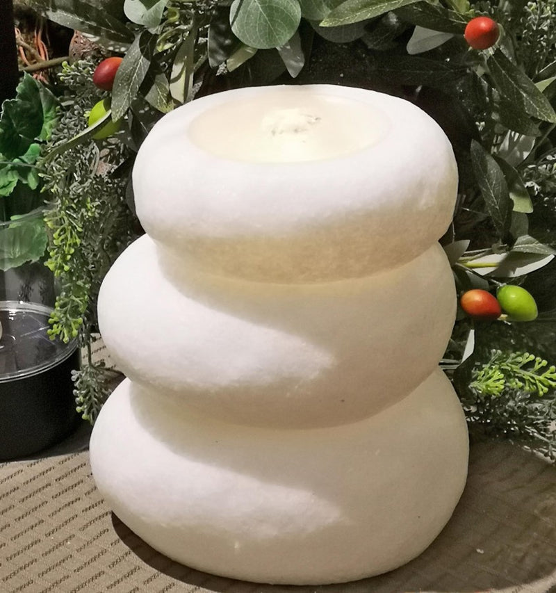 A three-tiered snowman-shaped white stone candle holder or fountain with a lit candle inside, set against a background of green foliage and small red and green berries.