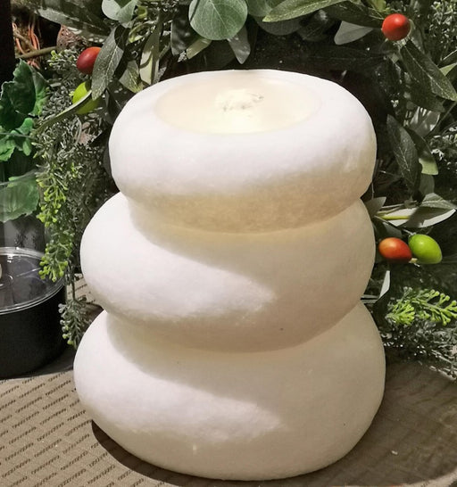 A three-tiered snowman-shaped white stone candle holder or fountain with a lit candle inside, set against a background of green foliage and small red and green berries.