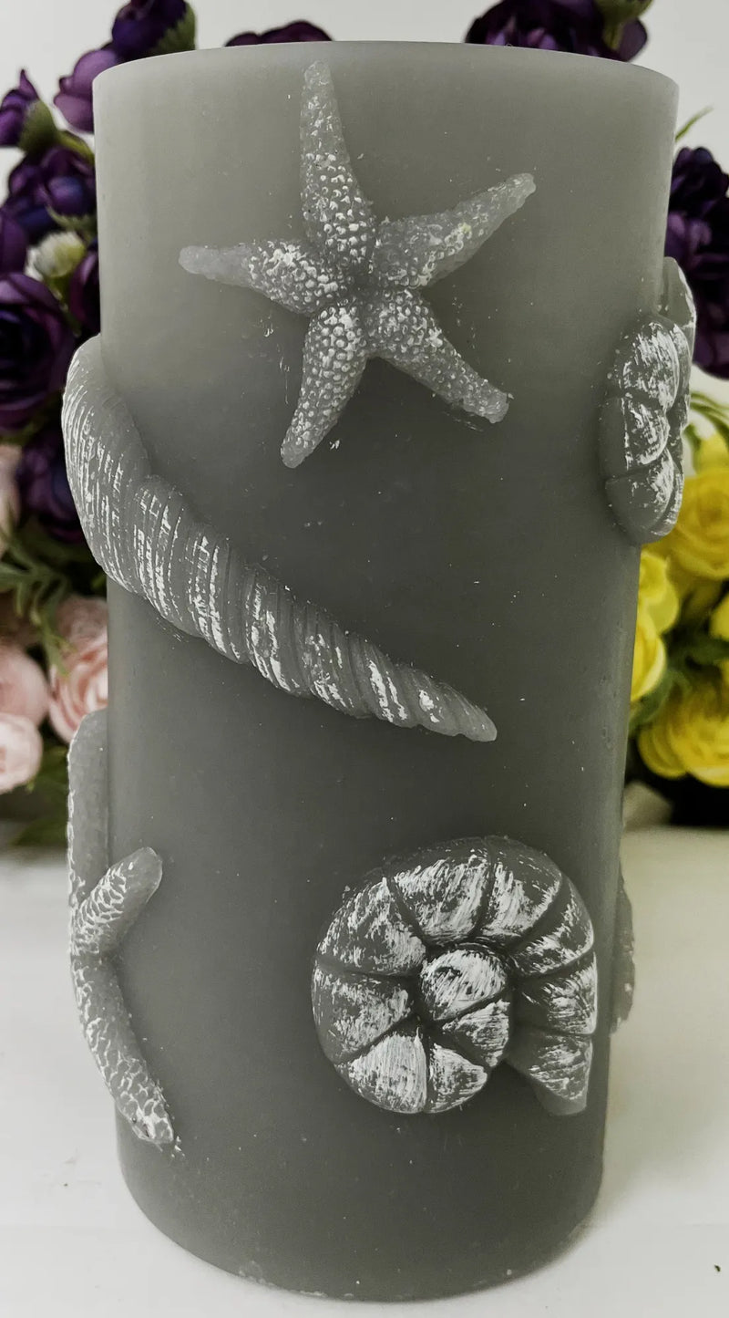 A dark gray cylindrical candle decorated with raised silver seashell and starfish motifs, shown against a blurred floral background.