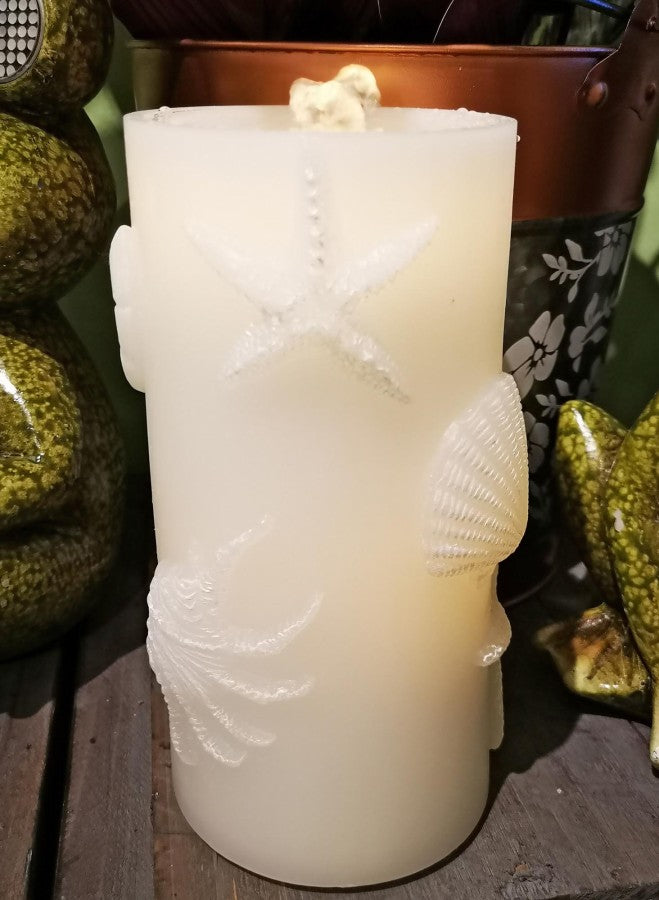 A tall, cylindrical cream-colored candle with embossed sea-themed motifs including a starfish, seashell, and starfish, displayed on a dark wooden surface with decorative frog figurines in the background.