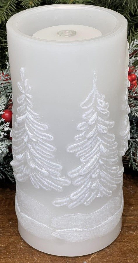 A frosted white pillar candle with embossed Christmas tree designs, shown on a wooden surface with holiday greenery and red berries in the background.