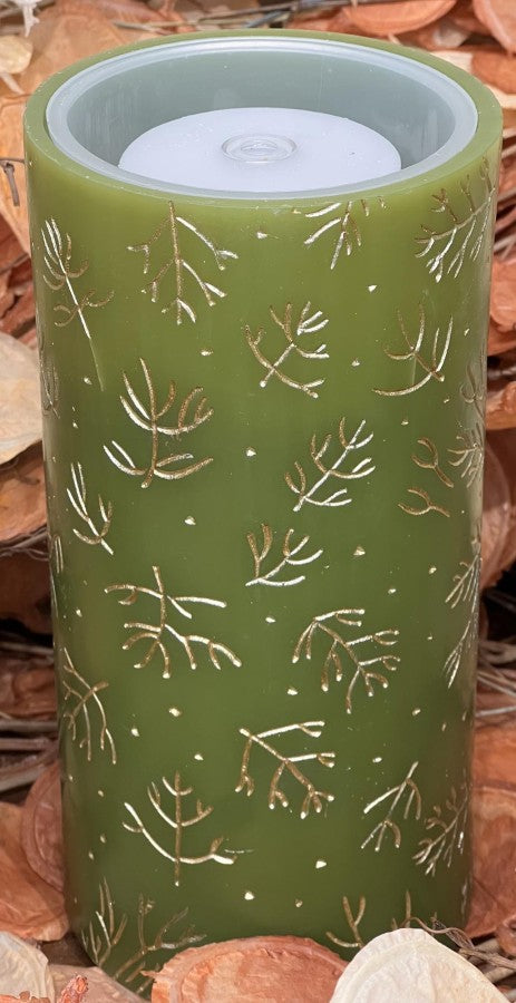 An olive green cylindrical candle with a white inner cup, decorated with a pattern of gold-toned pine branches and small white dots, set against a background of dried autumn leaves.