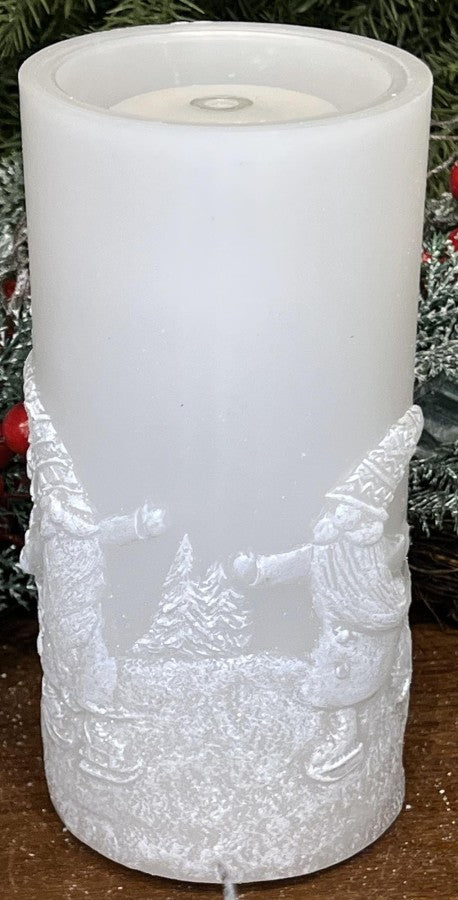 A white pillar candle with a frosted finish and embossed holiday figures at the base, shown standing upright against a festive background of green pine branches and red ornaments.