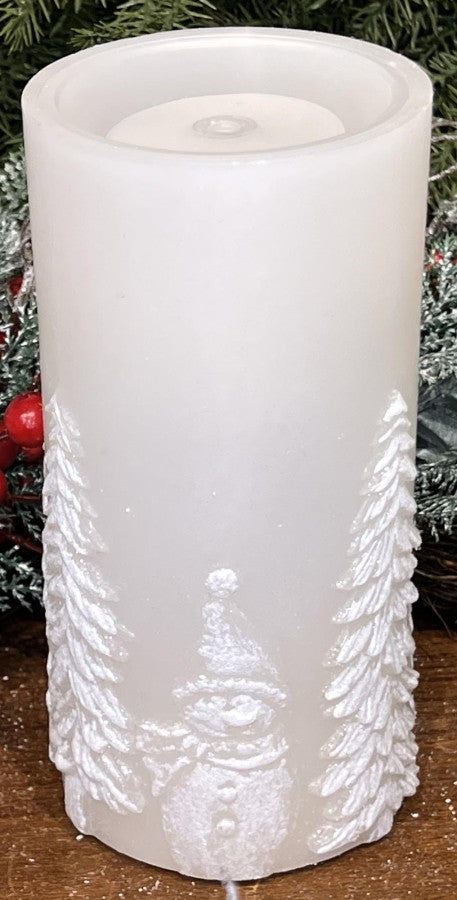 A white pillar candle with embossed Christmas tree and gnome designs, shown on a wooden surface with holiday greenery and red berries in the background.