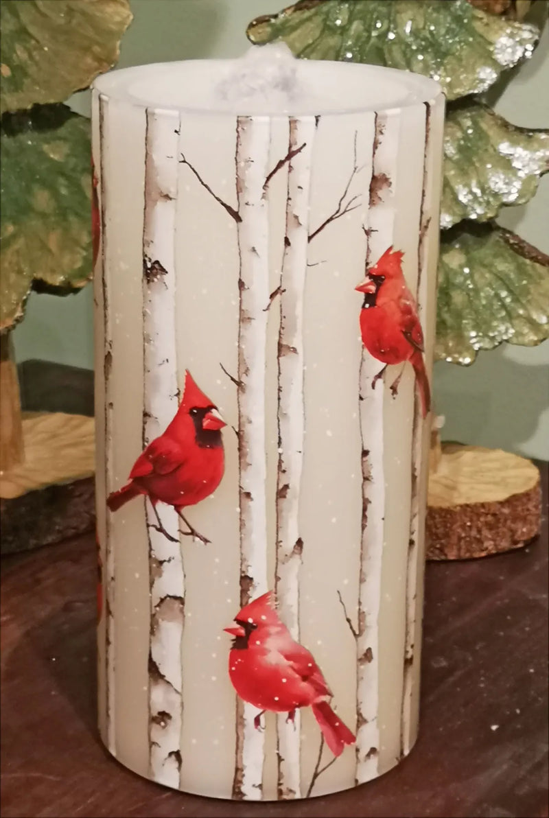 A cylindrical decorative candle with a birch tree bark pattern and three red cardinal birds printed on the front, shown on a tabletop with holiday greenery in the background.