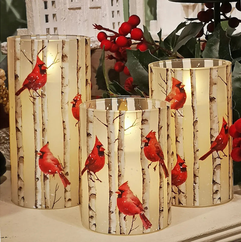Three cylindrical glass candle holders with a mirrored birch tree pattern and red cardinal bird designs, lit with warm internal lights, set against a holiday background with red berries and green leaves.