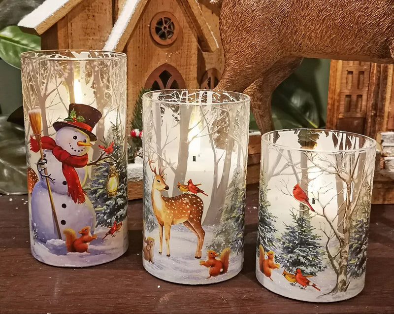 Three tall clear glass candle holders with winter holiday illustrations, including a snowman, deer, and cardinals in snowy forests, displayed on a wooden surface with a rustic house backdrop.