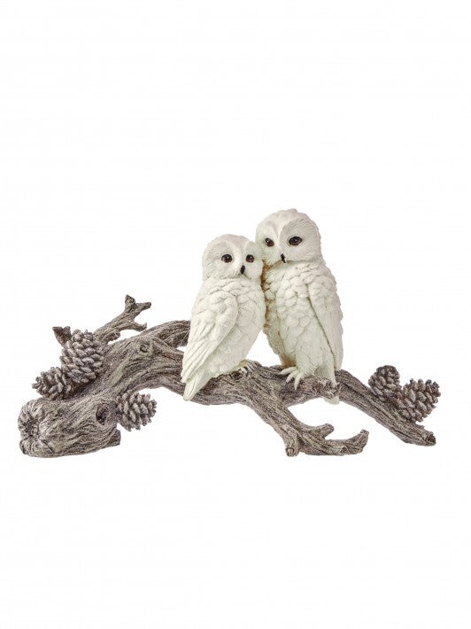 A decorative figurine of two white owls perched on a weathered gray branch with small pinecones, isolated on a white background.