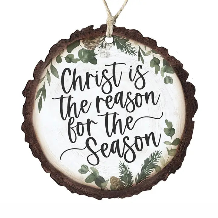 A round hanging Christmas ornament shaped like a rustic tree slice with a dark brown bark edge, a white center, black script text reading 'Christ is the reason for the Season,' and green botanical illustrations around the border. A natural twine loop hangs from the top.