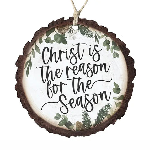 A round hanging Christmas ornament shaped like a rustic tree slice with a dark brown bark edge, a white center, black script text reading 'Christ is the reason for the Season,' and green botanical illustrations around the border. A natural twine loop hangs from the top.