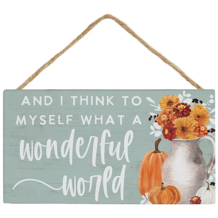 A rectangular hanging wooden sign with a rustic twine handle. The sign has a distressed light blue-green background with white text reading 'AND I THINK TO MYSELF WHAT A wonderful world' and an autumn floral illustration featuring orange pumpkins, a white pitcher, and yellow-orange flowers on the right side.
