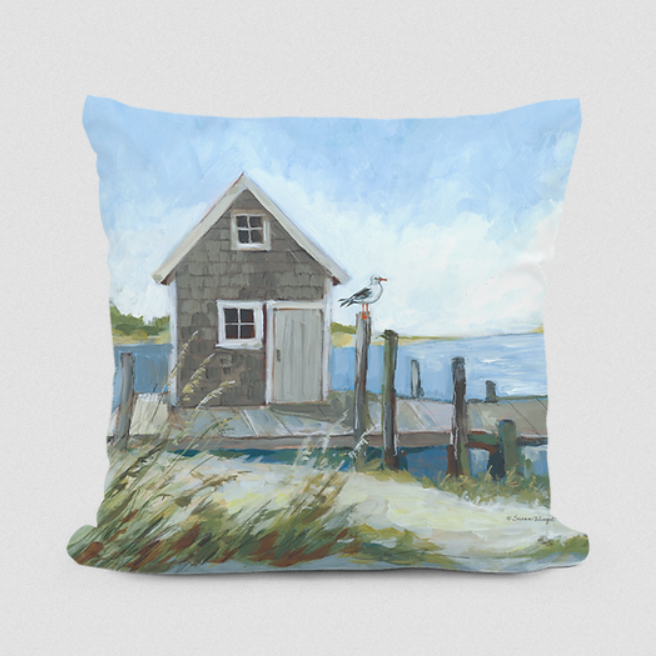 A decorative pillow with an artistic painting of a boat house on a pier, with a bird perched on the fence, under a blue sky with clouds.