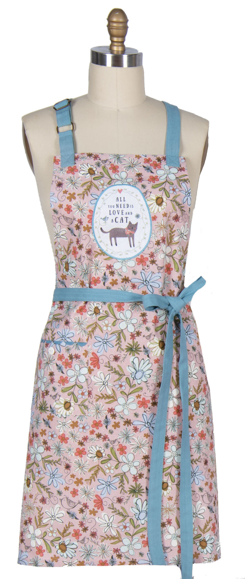 A floral apron with light blue straps and waist tie, displayed on a cream dress form mannequin. The apron features a multicolor floral pattern on a light pink background with a central circular patch containing a moose illustration and the text 'ALL THAT YOU LOVE AND A CAT'.