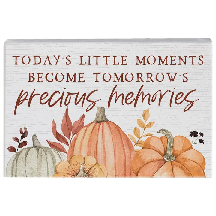 A rectangular decorative sign with a white wood-textured background and autumn watercolor pumpkins along the bottom. The sign reads, 'TODAY'S LITTLE MOMENTS BECOME TOMORROW'S precious memories' in brown lettering.