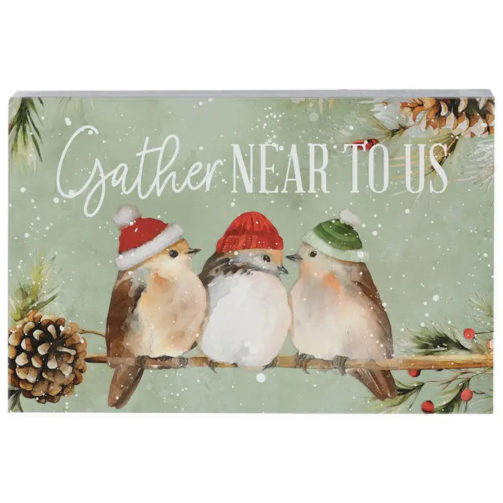 A square decorative sign or print featuring three small birds perched on a branch, each wearing a festive Santa hat, with the phrase “Gather Near to Us” in white script across a snowy mint-green background. Pinecones, evergreen branches, and red berries decorate the scene.