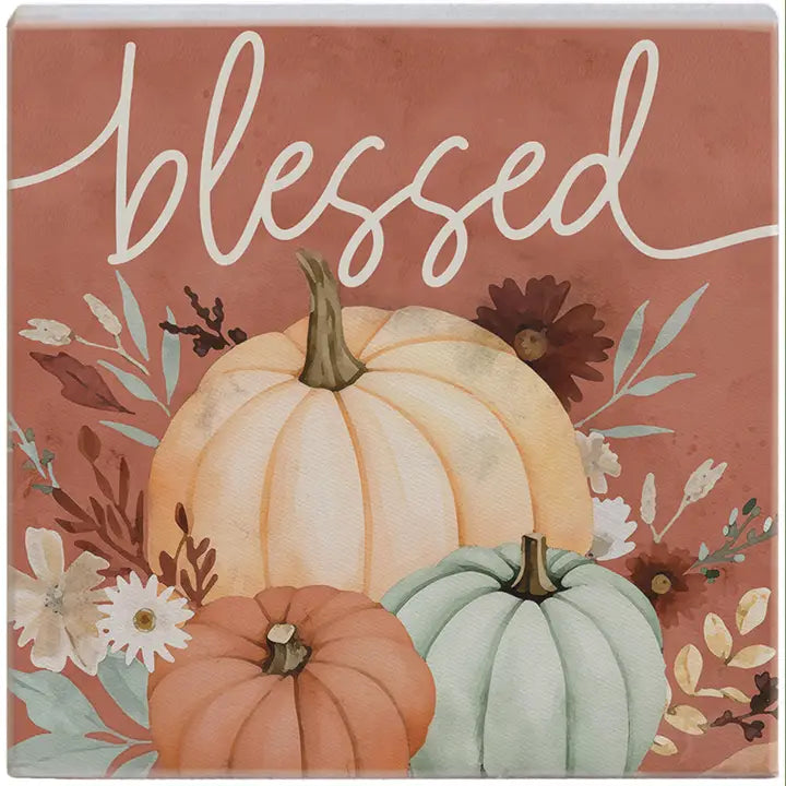Square decorative sign with the word 'blessed' in white script over a watercolor autumn design featuring two pumpkins, white daisies, and fall leaves on a dusty terracotta background.