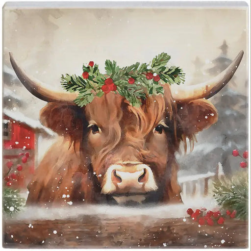 A square decorative artwork featuring a festive watercolor illustration of a brown Highland cow with long horns, wearing a green holly wreath with red berries. The background depicts a snowy, softly blurred winter scene with a red barn and distant house.