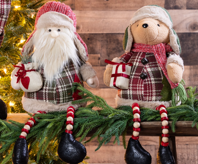 Two decorative figures, one resembling Santa Claus and the other a snowman, sitting on a wooden surface with a Christmas tree in the background.