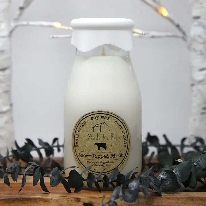 Milk bottle candle labeled 'Snow-Tipped Birch' on a decorative surface with greenery.