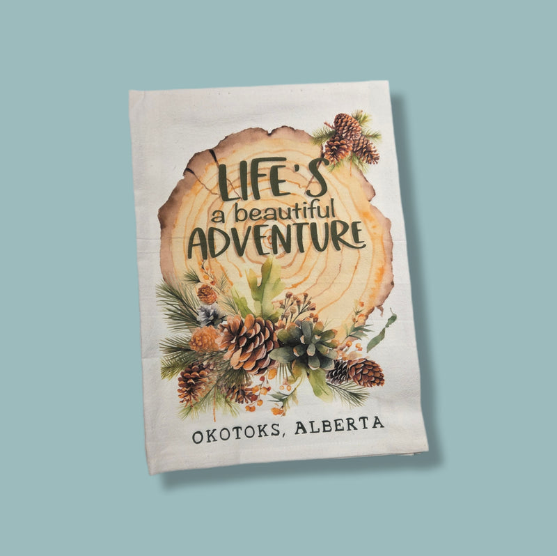 Decorative towel with 'Life's a Beautiful Adventure' text and pinecones on a light blue background
