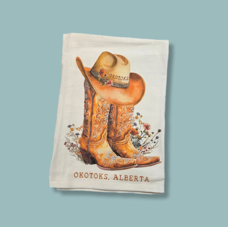 Tea towel with cowboy boots and hat design featuring 'Okotoks, Alberta' text on a light blue background