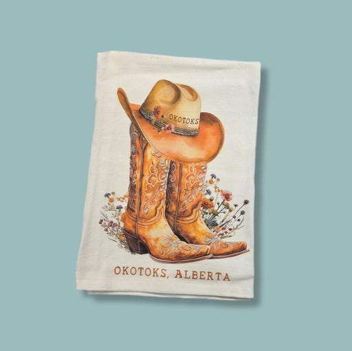Tea towel with cowboy boots and hat design featuring 'Okotoks, Alberta' text on a light blue background
