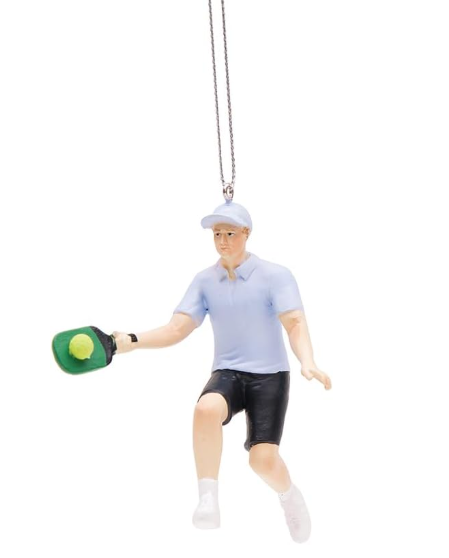 A resin ornament in the shape of a pickleball player wearing a blue shirt and black shorts, holding a green pickleball and a paddle, with a cap on the head, hanging by a string.