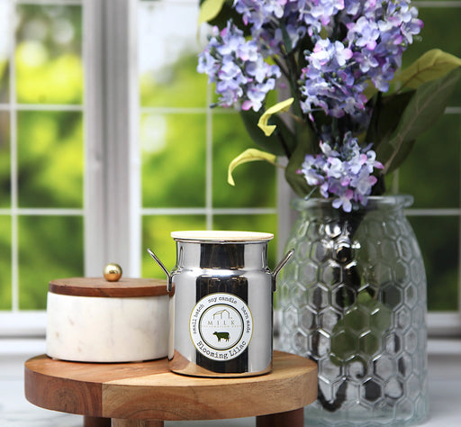 Decorative milk churn and glass vase with flowers on a wooden stand by a window.