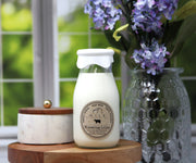 Glass bottle candle labeled 'Milk' with a floral arrangement in the background