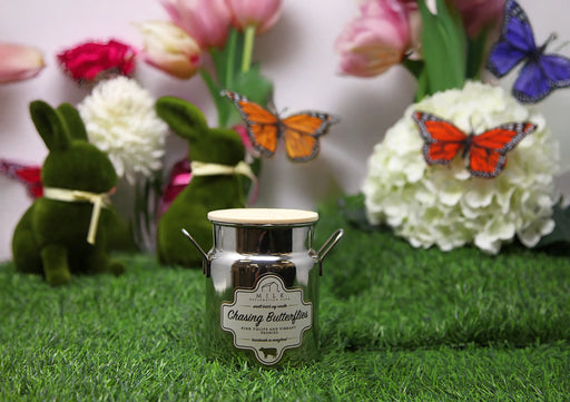 Candle with 'Chasing Butterflies' label on grass with decorative elements like flowers, butterflies, and a rabbit.