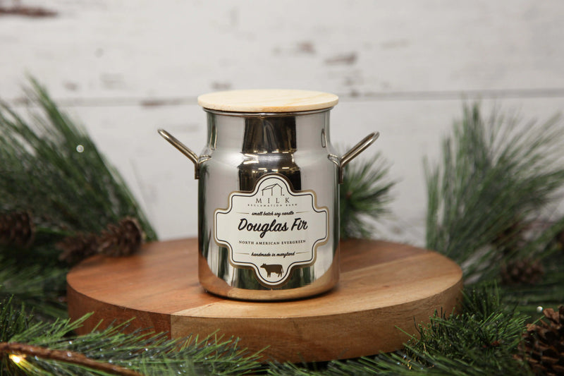 Candle in a metal container with a label on a wooden stand surrounded by greenery