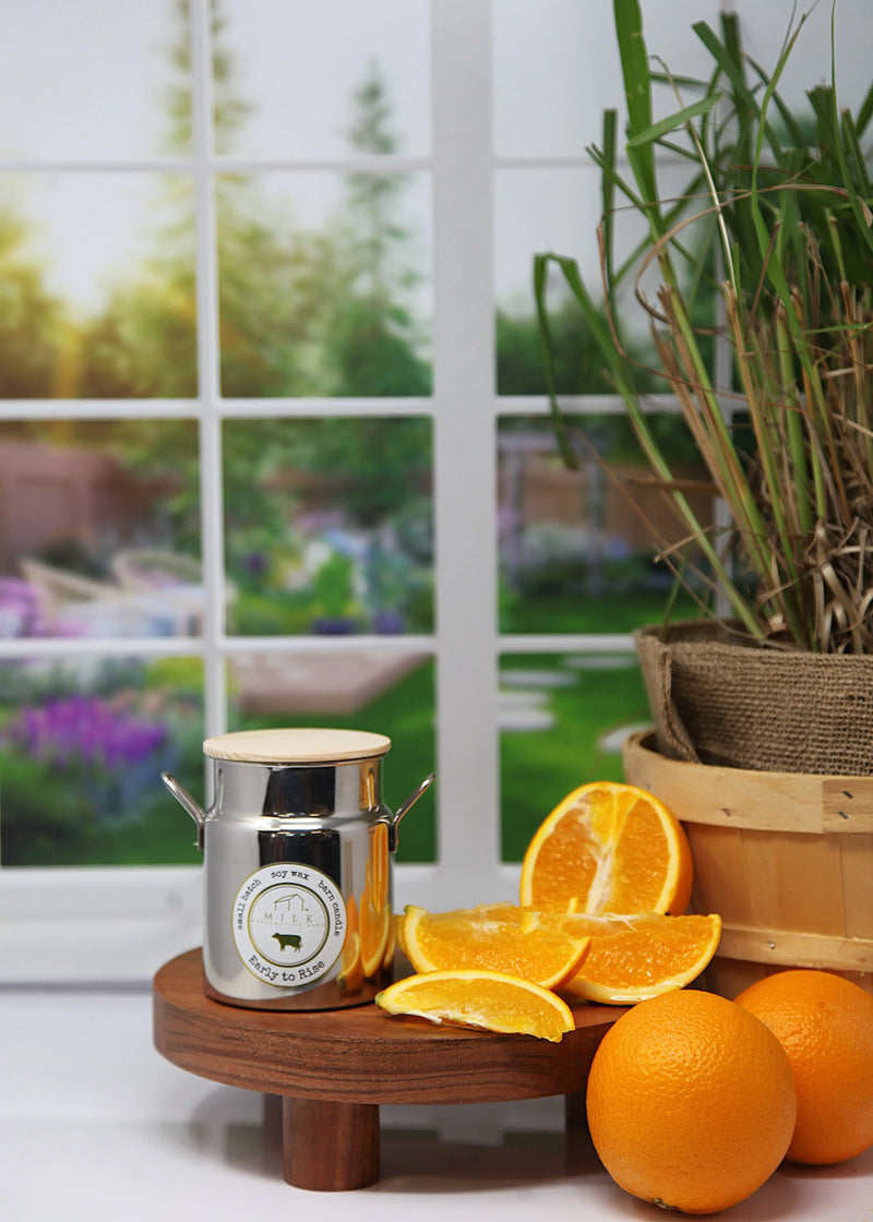 Silver candle with a label on a wooden stand with sliced oranges and a plant in the background.