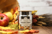Candle labeled 'Autumn Leaves' in a decorative pot with fall elements on a wooden surface.