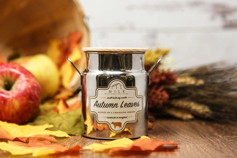 Candle labeled 'Autumn Leaves' in a decorative pot with fall elements on a wooden surface.
