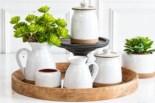 A styled tabletop arrangement of white ceramic pitchers, jars, and a small planter with green succulent plants, set on a round wooden tray against a white paneled wall.