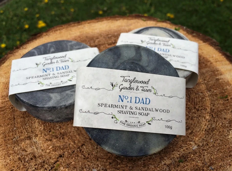 Three Tanglewood Garden & Farm shea mint and sandalwood shaving soaps on a wooden block with grass in the background.