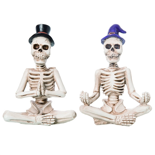 Two skeleton figurines in meditation poses with hats on a white background